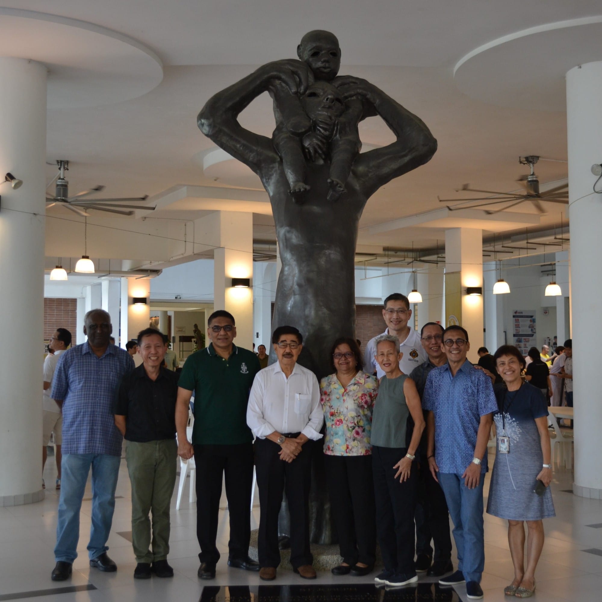 Ten people stand posing in front of a tall dark bronze sculpture of a figure holding a child indoors.